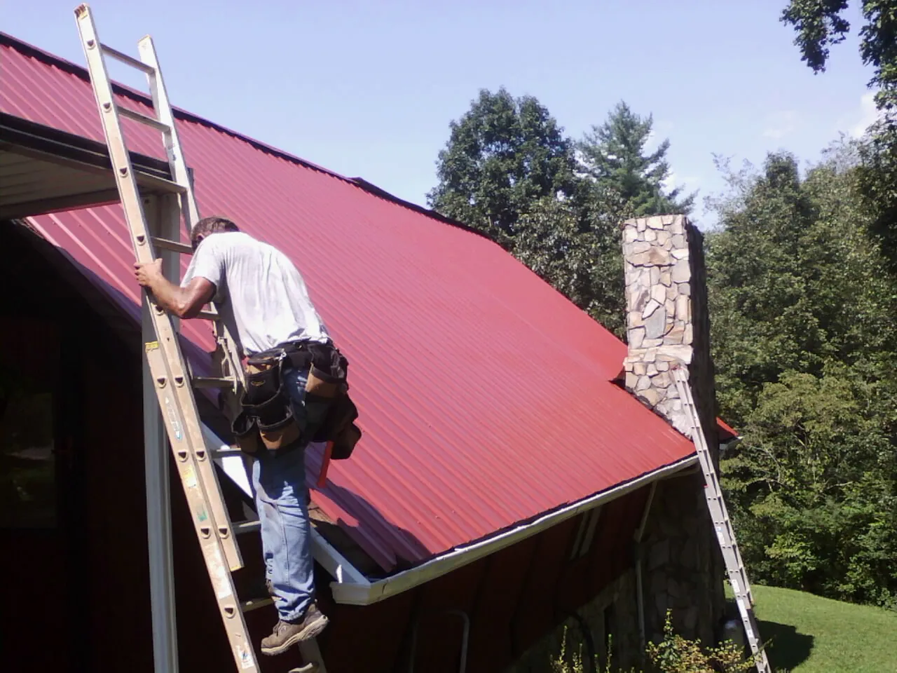 Roofer on a ladder beside a red metal roof and stone chimney — Soffit & Fascia Repair by Roofing Brothers in Granite Springs